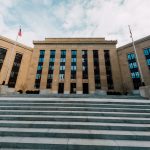 Fachada do edifício do Federal Reserve, vista de baixo, com bandeiras dos Estados Unidos e céu nublado.