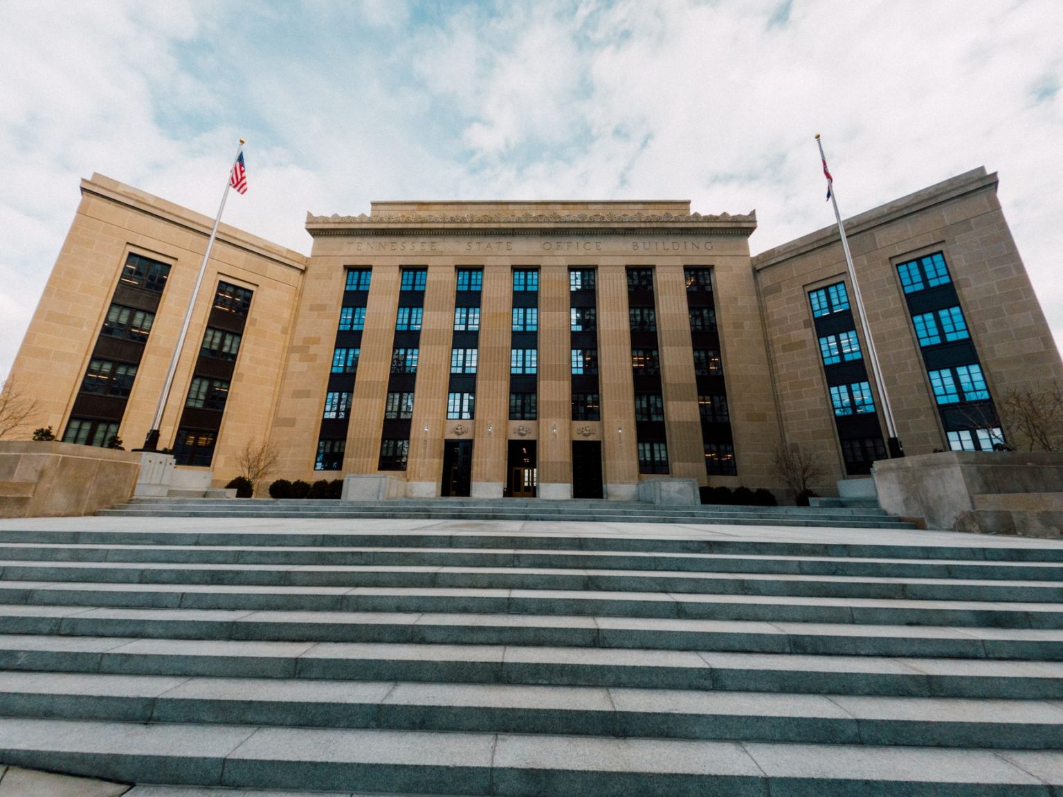Fachada do edifício do Federal Reserve, vista de baixo, com bandeiras dos Estados Unidos e céu nublado.