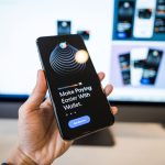 Mão segurando um smartphone com a interface de uma carteira digital, diante de um monitor desfocado.