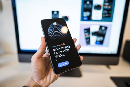 Mão segurando um smartphone com a interface de uma carteira digital, diante de um monitor desfocado.