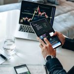 Pessoa analisando gráficos financeiros em um tablet e um laptop, em ambiente de análise de mercados digitais.