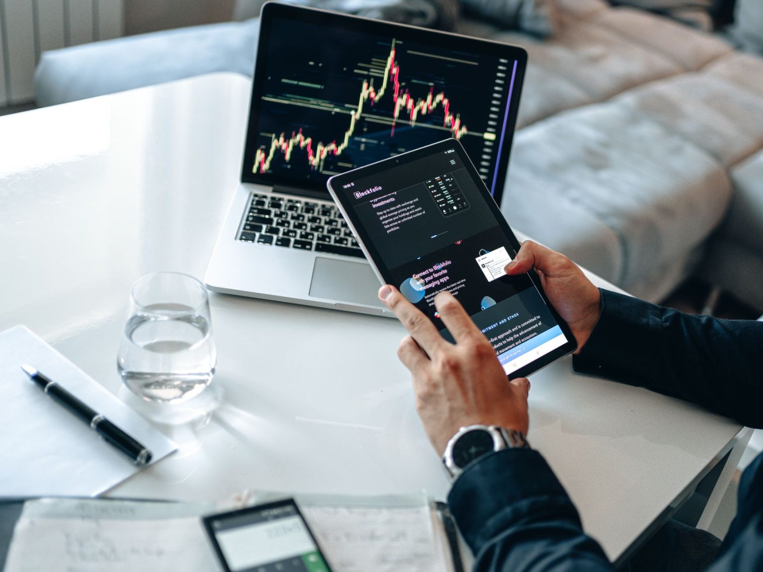 Pessoa analisando gráficos financeiros em um tablet e um laptop, em ambiente de análise de mercados digitais.