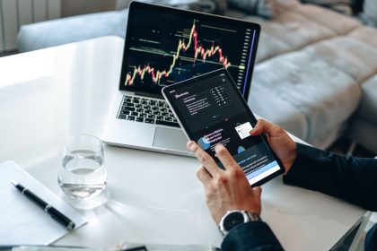 Pessoa analisando gráficos financeiros em um tablet e um laptop, em ambiente de análise de mercados digitais.