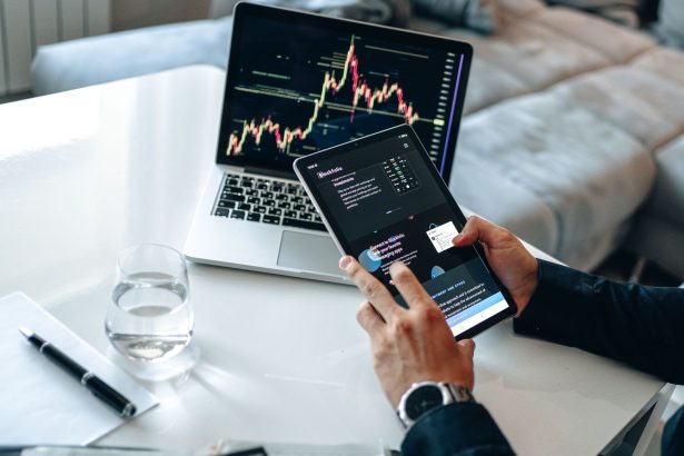 Pessoa analisando gráficos financeiros em um tablet e um laptop, em ambiente de análise de mercados digitais.
