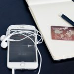 Smartphone branco, cartão de crédito, fones de ouvido e caneta sobre um bloco de anotações, compondo uma cena relacionada a tecnologia e pagamentos.