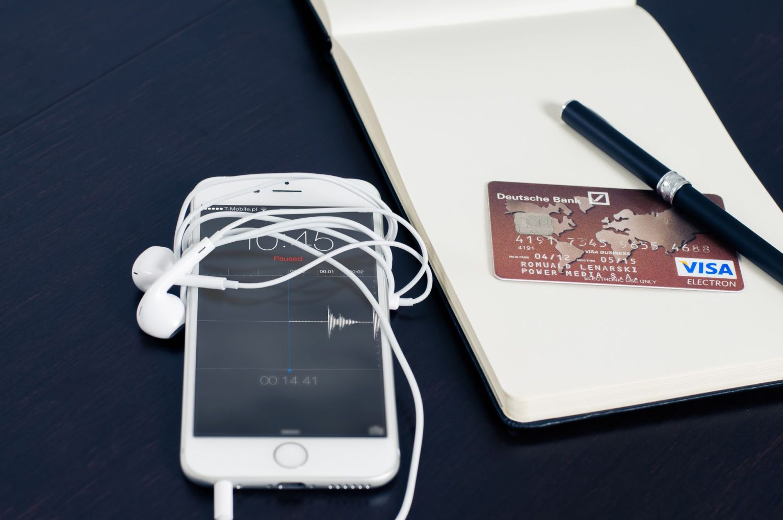 Smartphone branco, cartão de crédito, fones de ouvido e caneta sobre um bloco de anotações, compondo uma cena relacionada a tecnologia e pagamentos.