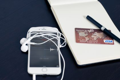 Smartphone branco, cartão de crédito, fones de ouvido e caneta sobre um bloco de anotações, compondo uma cena relacionada a tecnologia e pagamentos.