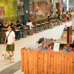 Pessoas circulam por um shopping movimentado, com lojas e área de alimentação ao fundo.